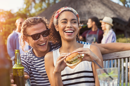 Cheerful multiethnic couple enjoying hamburger and beer at barbecue party. Happy young black woman eating burger while is sitting on boyfriend legs.の写真素材