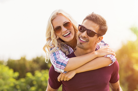 Closeup of young man carrying beautiful woman on his back.Â Smiling girl piggyback on guy while looking at camera. Boyfriend giving girlfriend piggyback ride and smiling outdoor.の写真素材