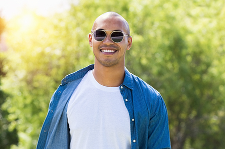 Portrait of smiling african man looking at camera with sunglasses. Happy guy enjoying the summer outdoor. Cheerful young man wearing white tshirt and blue shirt at park.の写真素材