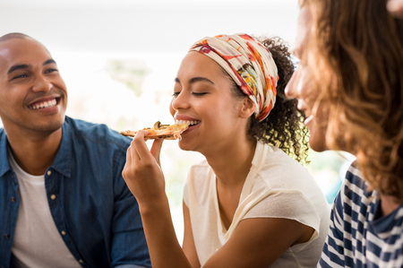 Portrait of african woman sitting with friends eating pizza slice. Friends having pizza party at home. Men and young woman having fun together at home during lunch break.の写真素材