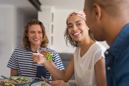 Beautiful african woman eating fresh salad with friends for lunch. Happy cheerful friends in a conversation during lunch at home. Young friends meeting each other for healthy meal.の写真素材