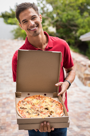 Young delivery man in red uniform holding a pizza. Portrait of happy delivery boy showing pizza in a cardboard box.の写真素材