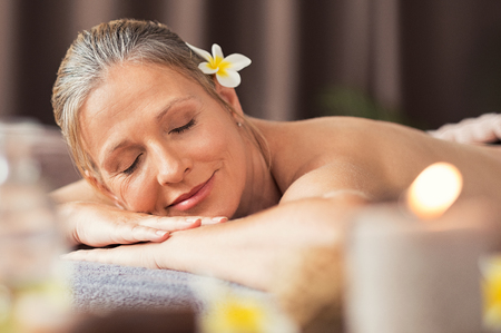 Beautiful blond woman relaxing at spa after body massage. Portrait of mature woman lying on massage table with closed eyes. Senior woman lying on a lounger at wellness center with candles.の写真素材