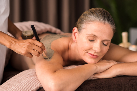 Relaxed mature woman enjoying a mud skin treatment at spa. Portrait of beautiful senior woman having clay body mask applyed by beautician in a wellness center. Exfoliation scrub beauty treatment.の写真素材