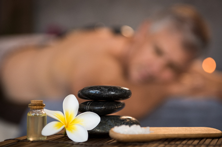 Closeup of stacked black hot stones on wooden table with oil, rock salt and frangipani flower. Zen stones in spa center with cosmetics and massage products. Wellness, beauty and wellbeing concept.の写真素材