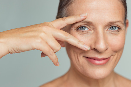 Portrait of smiling senior woman with perfect skin showing victory sign near eye on grey background.  Closeup face of mature woman showing successful results after anti-aging wrinkle treatment. Beauty mature skin care concept.