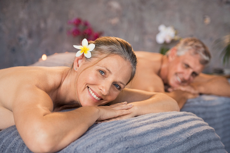 Senior man and beautiful woman lying down on massage beds at spa. Mature couple lying on massage table and smiling while looking at camera. Happy married couple relaxing at wellness spa resort.の写真素材