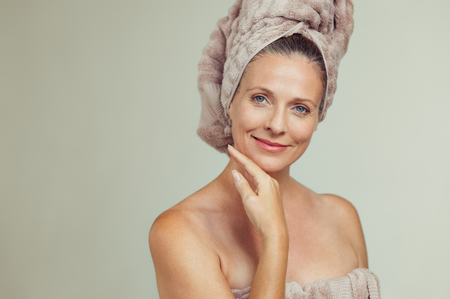 Portrait of smiling senior woman feeling soft skin with hand on chin after spa bath. Mature woman draped in towel on body and wet hair looking at camera. Lady in bathrobe after shower isolated on grey background.の写真素材