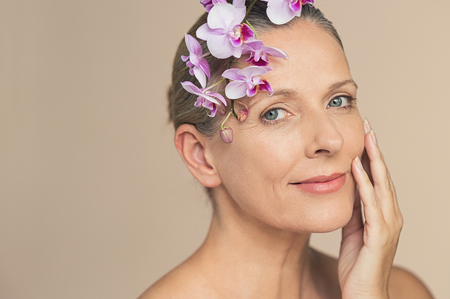 Portrait of beautiful senior woman looking at camera with a flower on head. Closeup face of beauty mature woman holding purple orchid isolated on grey background. Mature skin face with wrikles eyes.の写真素材