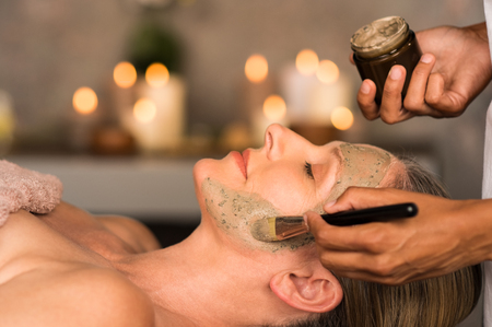 Senior woman relaxing while beautician getting purifying clay mask on her face. Closeup hand of masseuse applying mud from jar container on face of mature lady in spa center. Anti aging facial treatment with brush.の写真素材