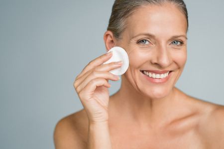 Healthy mature woman removing makeup from her face with cotton pad isolated on grey background. Beauty portrait of happy woman cleaning skin and looking at camera.