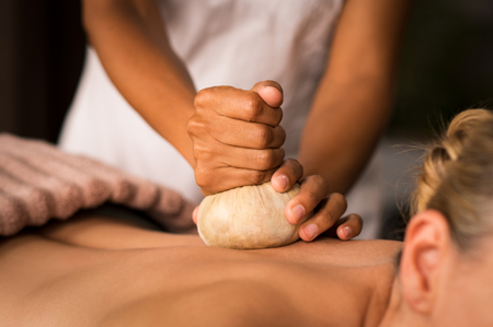 Closeup of masseuse hands holding cloth giving pinda massage to woman on spa table. Therapist hands giving ayurveda aroma pinda sweda massage to naked lady. Senior woman doing aromatic painkiller massage on back.の写真素材