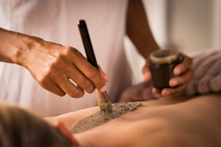 Closeup of beautician applying clay with brush on back of woman lying on massage table at spa. Woman hands using brush for clay mask. Beautiful lady receiving a scrub exfoliation therapy on body in a wellness center.の写真素材