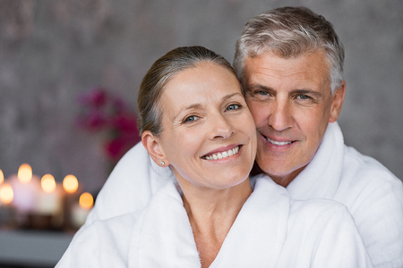 Portrait of mature married couple embracing with bathrobe and looking at camera. Happy senior man and beautiful woman in robes smiling at wellness center. Man and wife relaxing together at day spa.の写真素材