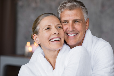 Smiling husband embracing cheerful wife from behind at spa. Laughing mature couple enjoying a romantic hug at wellness center after massage. Senior man and woman in white in bathrobe relaxing at spa.の写真素材