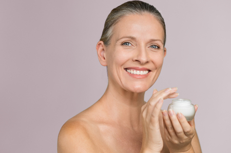 Beautiful mature woman holding jar of moisturizer and looking at camera. Happy senior woman holding a bottle of anti-aging lotion isolated over grey background. Anti aging and beauty treatment.の写真素材