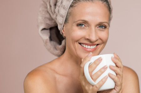 Portrait of mature smiling woman with hair covered in towel holding tea mug against grey background. Senior woman sipping infusion after shower. Happy lady with hair wrapped in towel enjoying hot beverage after spa treatment.の写真素材