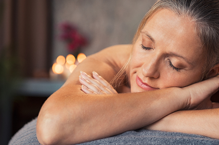 Closeup face of mature woman lying on a massage table with eyes closed in a wellness center. Blonde woman lying on front and relaxing after beauty massage. Portrait of serene senior woman awating massaging in a spa salon.の写真素材