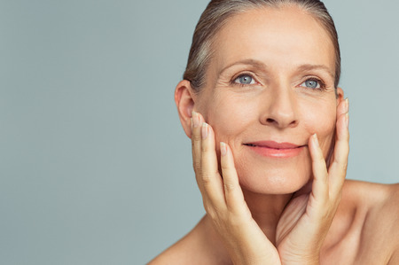 Portrait of mature woman with perfect skin isolated on grey background. Closeup face of happy senior woman with hands on cheeks looking away. Facing aging with a carefree attitude.