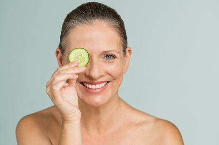 Portrait of smiling senior woman covering an eye with a cucumber slice isolated on grey background. Beauty mature woman holding slice of cucumber and looking at camera. Skincare and facial treatment.