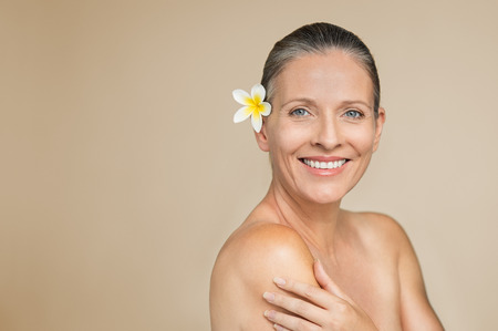 Happy mature woman with frangipani flower in hair smiling isolated over grey background. Beautiful middle aged woman with naked shoulder looking at camera with copy space. Portrait of senior beauty after skin care treatment.の写真素材