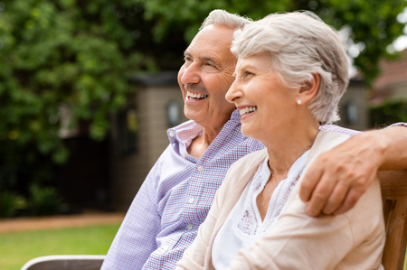 Senior couple sitting together on bench at park. Elderly married couple sitting outdoor and relaxing. Romantic husband embrace his wife while looking away and smiling. Future and retirement concept.の写真素材