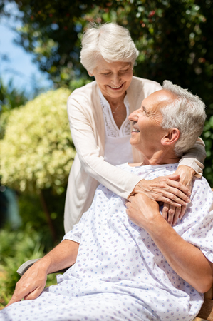 Senior woman hugging man in medical cloth in the hospital garden. Loving wife embracing old hospitalized husband sitting on bench at outdoor lawn. Caring wife supporting husband in illness in a private medical clinic.の写真素材