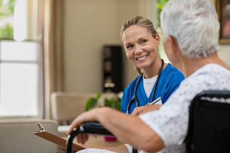 Friendly doctor examining health of patient sitting in wheelchair. Happy smiling nurse consulting disabled patient about treatment. Nurse caring about elder handicap man at home.の写真素材