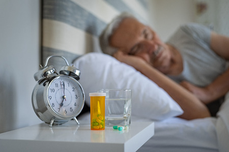 Medical pills and alarm clock with senior man sleeping in background. Silver alarm clock and medicines on table while man resting on bed. Bottle of capsules with glass of water kept on night table.の写真素材