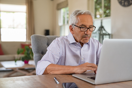 Senior man working with laptop at home. Old man using computer at home sitting on chair and looking at screen. Elderly grandfather wearing eyeglasses and working on laptop in living room.の写真素材