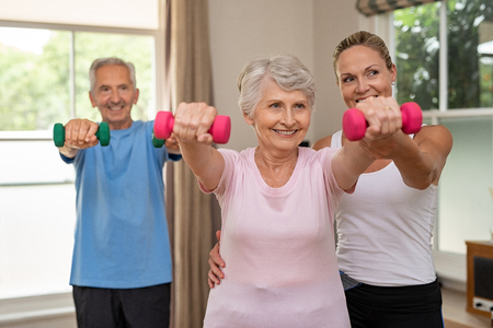 Happy senior couple exercising with dumbbells. Mature personal trainer helping elderly woman exercise at home. Old man and woman doing stretching exercise with physiotherapist.の写真素材