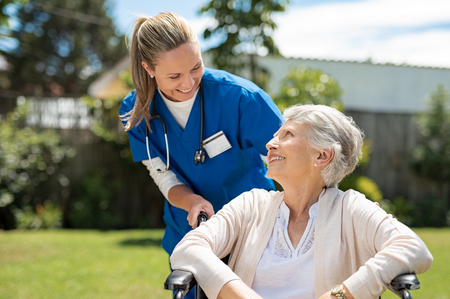 Nurse taking care of old woman in wheelchair outdoor. Friendly doctor caring about elderly disabled woman in wheelchair. Happy elderly woman with her caregiver at nursing home park.