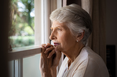 Depressed senior woman at home feeling sad. Elderly woman looks sadly outside the window. Depressed lonely lady standing alone and looking through the window.の写真素材