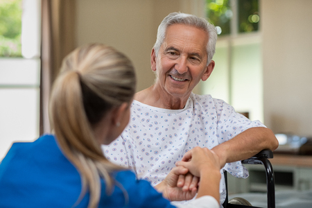 Smiling senior patient sitting on wheelchair with nurse holding hand. Old man sitting on wheelchair listening to doctor with a smiling face. Caring medical nurse comforting patient at home while holding hands.の写真素材