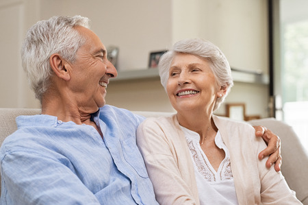 Romantic senior couple laughing while sitting on sofa. Elderly married couple smiling on sofa at home looking at each other. Happy old husband hugging his beautiful wife at home.の写真素材