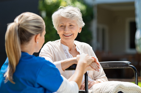 Smiling senior patient sitting on wheelchair with nurse supporting her. Doctor looking at elderly patient on a wheelchair in the garden. Nurse holding hand of mature woman outside pension home.の写真素材