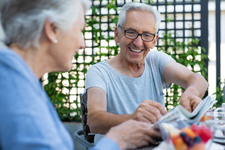 Elderly man wearing spectacles sitting with his wife for breakfast while he shows her a newspaper article. Portrait of a smiling senior man reading newspaper. Cheerful mature couple enjoying breakfast on balcony.の写真素材