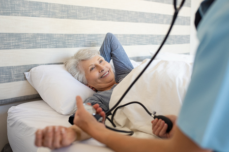 Nurse measuring blood pressure of senior woman with stethoscope and pressure machine at home. Happy mature woman lying on bed while getting blood pressure checked by doctor. Doctor taking care of old patient.の写真素材