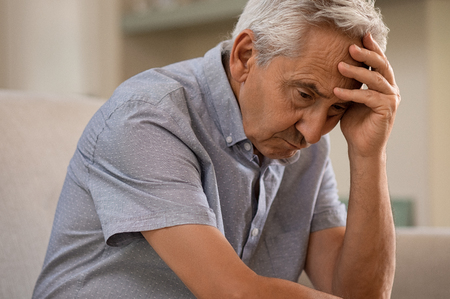 Thoughtful senior man sitting on couch. Depressed sad man sitting with hand on head thinking while looking away. Elderly man suffering from alzheimer.の写真素材