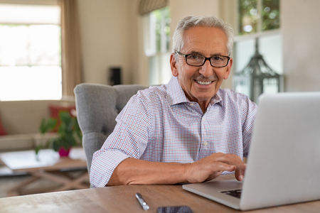 Smiling senior man looking at camera while using laptop at home. Handsome old man wearing eyeglasses working on laptop in living room. Portrait of elderly grandfather using computer.の写真素材