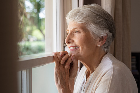 Happy senior woman standing at window and looking outside. Beautiful grandmother smiling while looking through the window. Portrait of cheerful old woman relaxing in home while standing at window sill.の写真素材