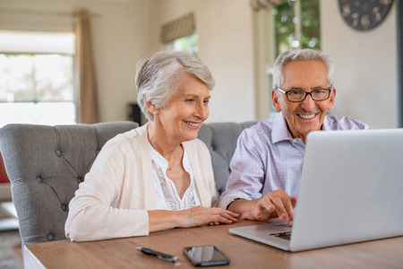 Happy smiling retired couple using laptop at home. Cheerful elderly man and old woman using computer while sitting at table. Smiling pensioner showing woman notebook at home.