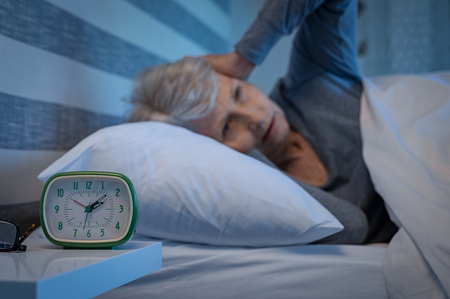 Old woman suffering from migraine unable to sleep during the late night. Elderly woman in bed ill and suffering from sleeplessness and insomnia. Closeup of alarm clock showing time with senior woman unable to sleep in background.の写真素材