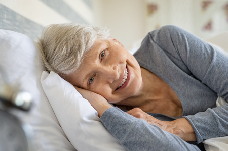 Smiling senior woman resting on bed and looking at camera. Awaken old woman with grey hair and pajamas in the early morning light. Portrait of elderly woman lying on side on bed and smiling.の写真素材