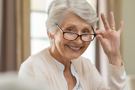 Happy retired senior woman looking at camera while holding eyeglasses. Smiling satisfied woman wearing spectacles at home. Closeup face of old grandmother trying on new eyewear.の写真素材