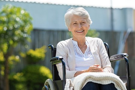 Happy senior woman sitting on wheelchair and recovering from illness. Handicapped mature woman sitting in wheelchair smiling and looking at camera. Portrait of a disabled elderly woman outdoor in a nursing home.の写真素材