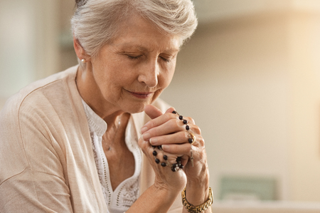 Senior woman praying while holding christian symbol of crucifix. Old woman praying to god with hope and closed eyes. Elderly believer make a prayer with faith holding rosary in hands.の写真素材