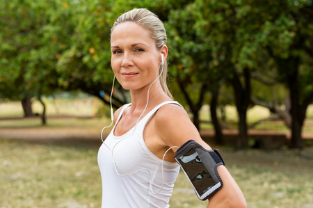 Portrait of athletic mature woman resting after jogging. Beautiful mid blonde woman running at park on a sunny day. Female runner listening to music while jogging and looking at camera.の写真素材