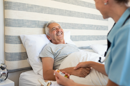 Cheerful senior man lying on bed receiving health care at home. Nurse advising old man on medication at home. Caregiver nurse helping elderly man taking medicine on the bedの写真素材