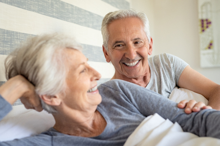 Smiling senior couple lying on bed on a bright sunny morning. Happy old man lay near his wife in bed. Mature couple having fun together in bedroom.の写真素材